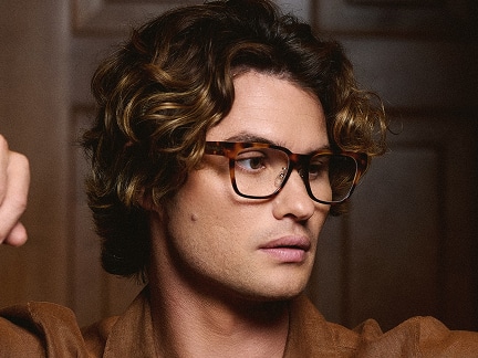 Chase Stokes wearing large, tortoiseshell glasses, looking slightly to the side with a neutral expression. He is dressed in a brown shirt, and the background features a softly focused wooden panel.