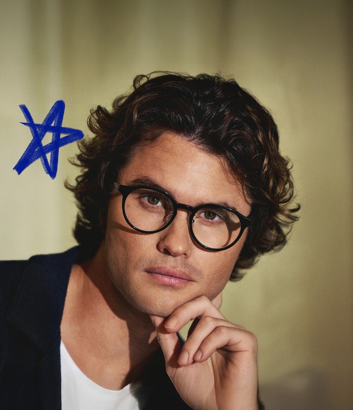 Chase Stokes is wearing round black Zenni glasses in front of a gold curtain backdrop with a hand-drawn blue star.