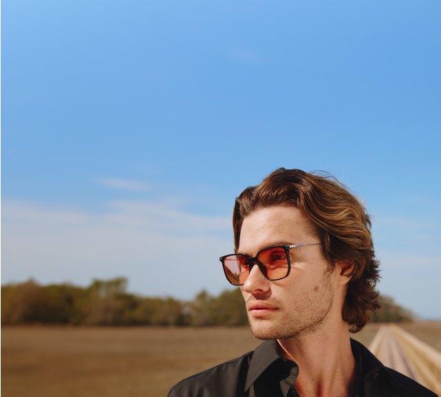 Chase Stokes wearing gradient tinted sunglasses and a black collared shirt looks off to the side against a scenic backdrop of an open road, clear blue sky, and distant greenery.