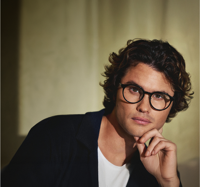 Chase Stokes is wearing round black Zenni glasses in front of a gold curtain backdrop with a hand-drawn blue star.