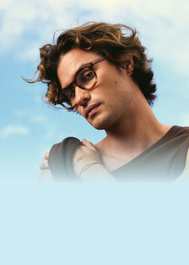Chase Stokes wears oversized brown Zenni eyeglasses against a blue, clouded sky.