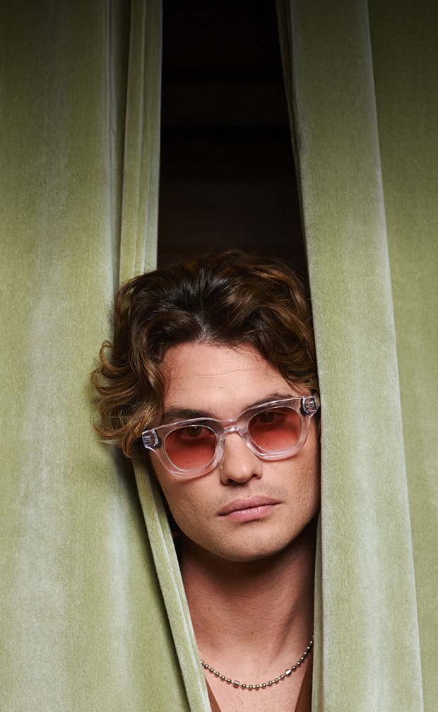 Translucent rectangle Zenni tinted glasses worn by Chase Stokes peeking through a green curtain.
