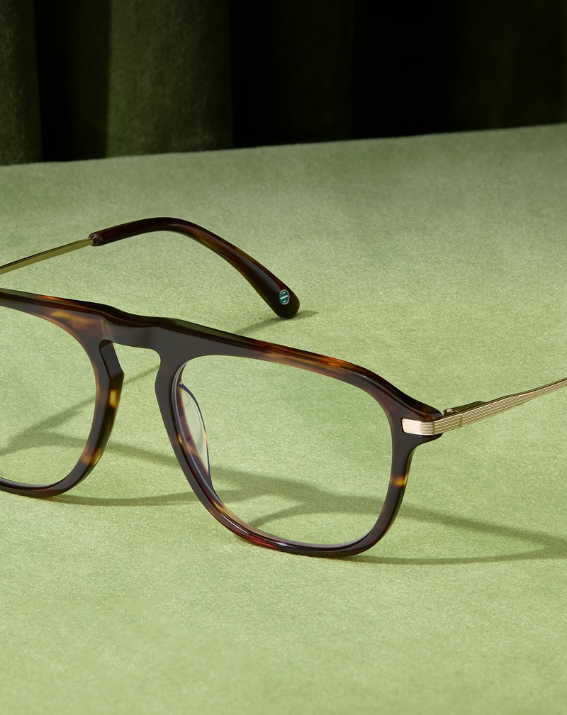 Dark tortoiseshell aviator eyeglasses with thin gold metal temples resting on a textured green surface.