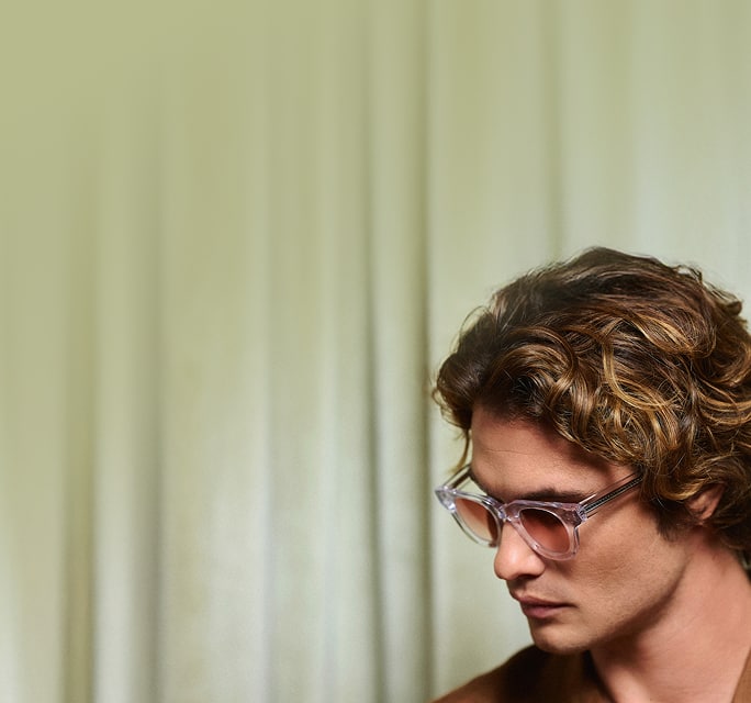 Chase Stokes looking downward in profile, wearing clear transparent acetate sunglasses with warm amber gradient lenses.