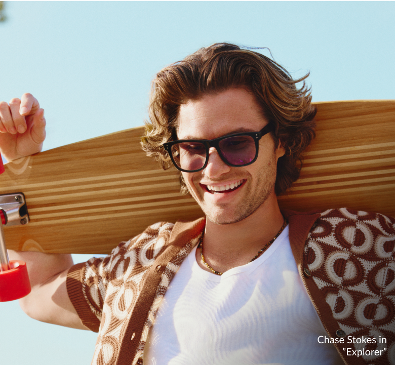 Chase Stokes in Zenni Jetsetter sunglasses, smiling outdoors in a white and brown geometric shirt against a blue sky with a wooden skateboard over his shoulders.