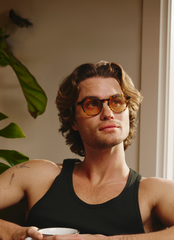 Chase Stokes sitting indoors holding a mug, wearing brown Zenni Surfwatch frames with tinted lenses and a black tank top