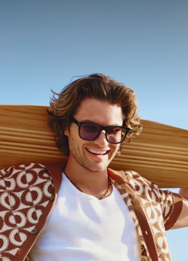 Chase Stokes in Zenni Jetsetter sunglasses, smiling outdoors in a white and brown geometric shirt against a blue sky with a wooden skateboard over his shoulders.