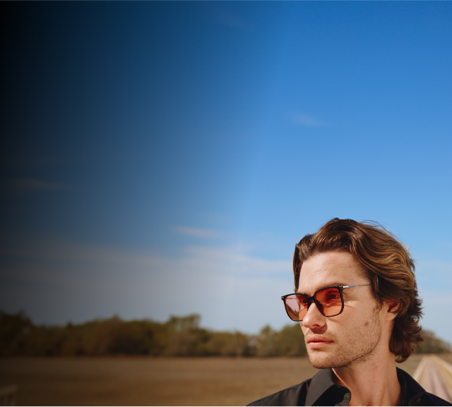 Chase Stokes is dressed in a black collared shirt, wearing square sunglasses with amber gradient tints. The background features a clear blue sky, an open field, and distant trees.