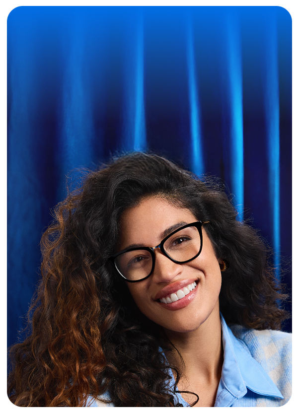 Woman wearing Zenni black square glasses and a light blue shirt, smiling against a deep blue curtain.