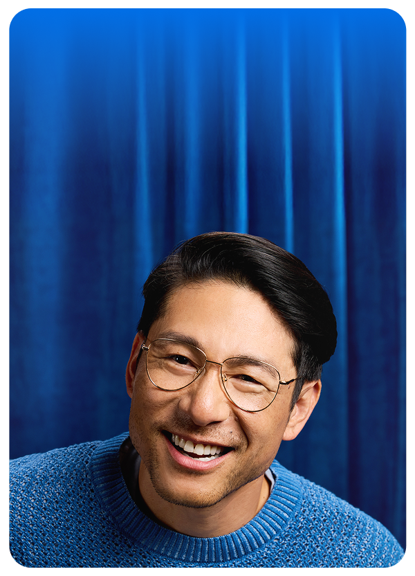 Man smiling in Zenni wireframe round glasses and a blue sweater, standing in front of a velvet blue curtain.