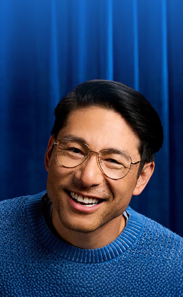 Man smiling in Zenni wireframe round glasses and a blue sweater, standing in front of a velvet blue curtain.
