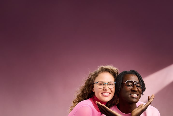 Two smiling people in pink clothing wearing Zenni glasses against a pink background.
