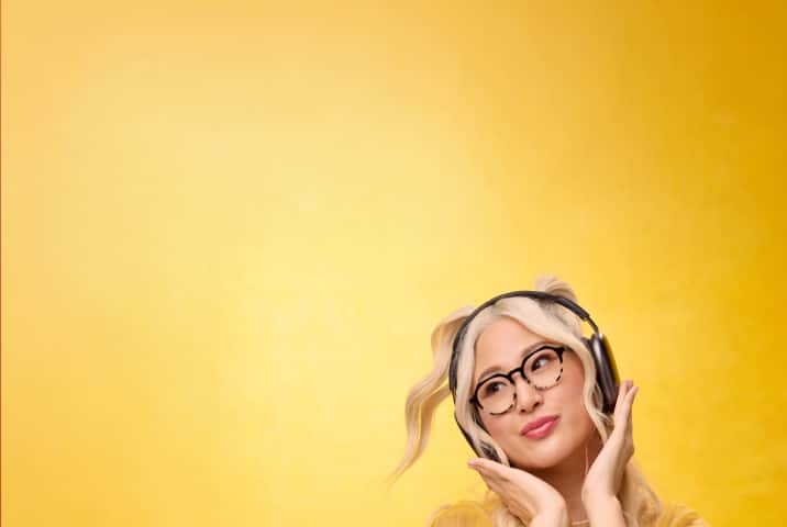 Blonde woman wearing round Zenni glasses, smiling with headphones on against a yellow background.