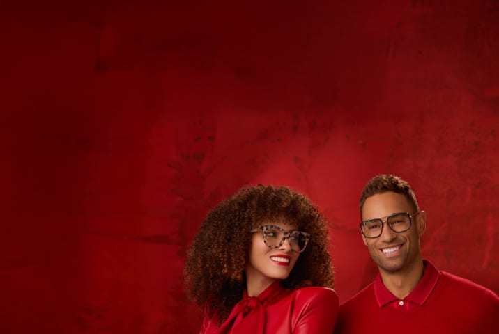 Two models in bold Zenni glasses posing against a rich red background - one woman with voluminous curls and a man in a red jacket.