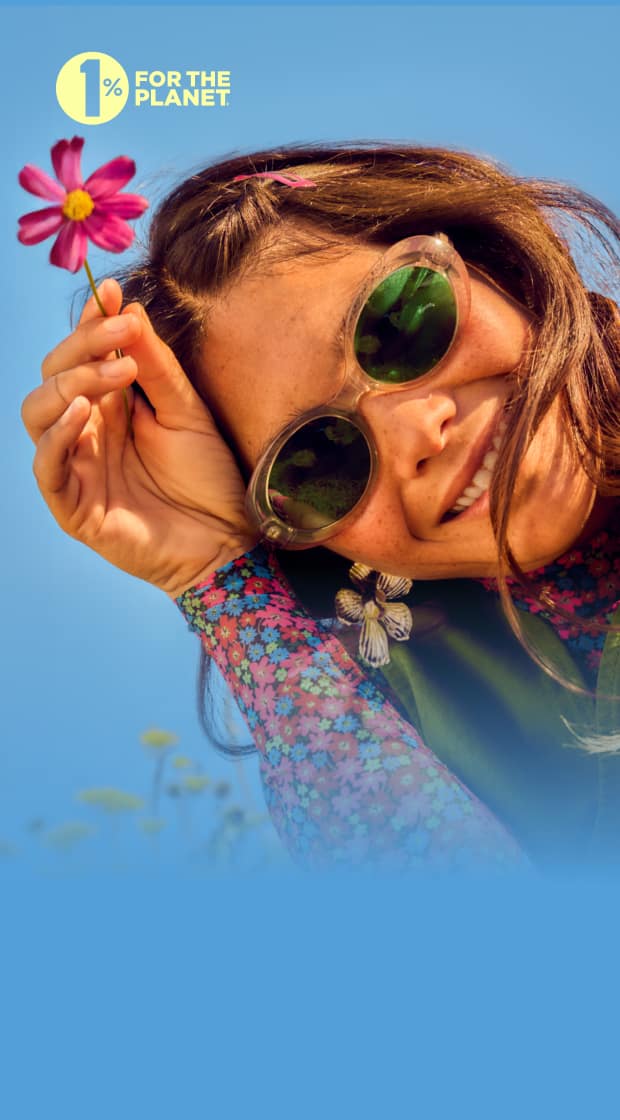 Smiling woman wearing translucent green sunglasses and a floral top, surrounded by blooming wildflowers, holding a pink daisy-like flower in her hand. ‘1% for the Planet’ badge in the corner.