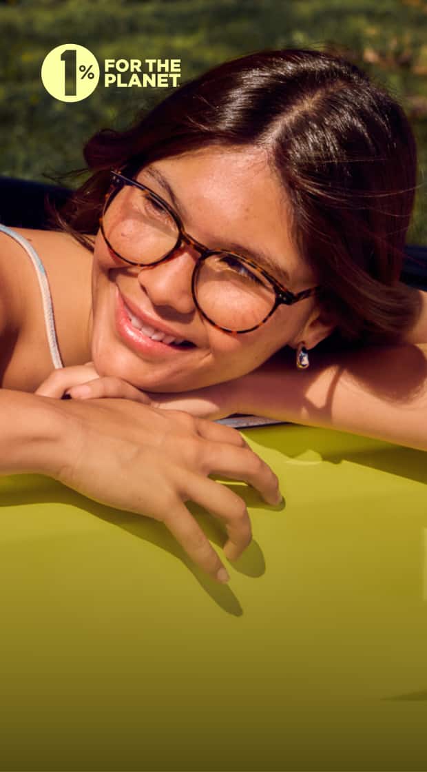 A Woman with medium brown hair and tortoiseshell glasses smiles while leaning on a bright yellow surface, with green foliage in the softly blurred background on a sunny day.