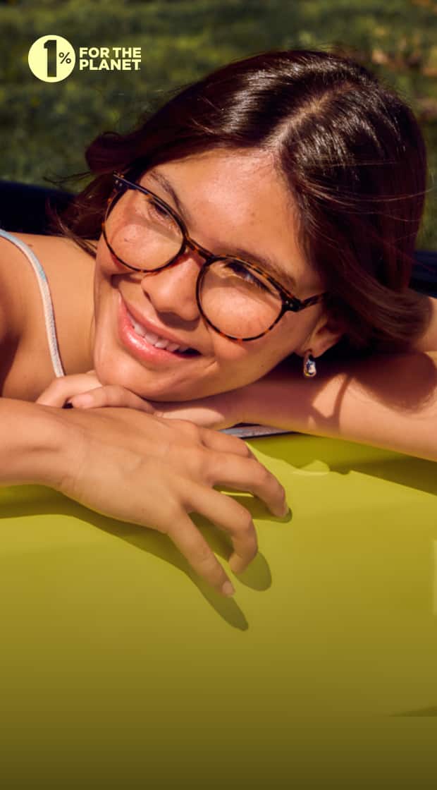 A woman with medium brown hair and tortoiseshell glasses smiles while leaning on a bright yellow surface, with green foliage in the softly blurred background on a sunny day.