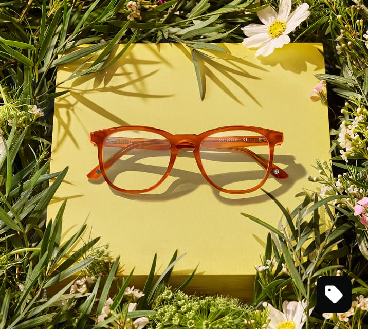 Amber EcoBloomz eyeglasses displayed on a yellow platform surrounded by grass and wildflowers, representing the eco-friendly eyewear collection.