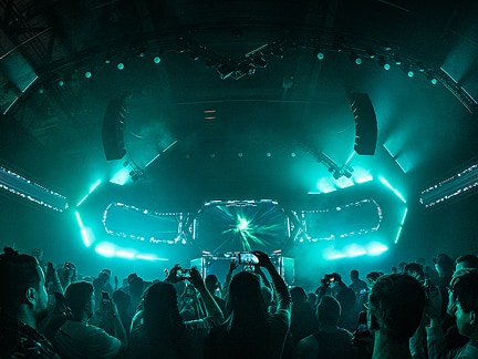 A lively concert scene with teal lighting shows a large crowd silhouetted before a bright stage, where beams and patterns of light create an energetic atmosphere.