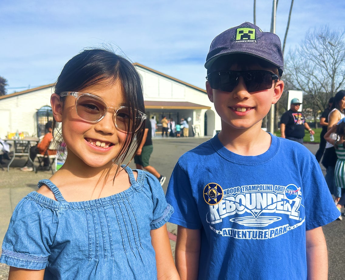 A young girl in a blue denim dress wearing clear, round-frame eyeglasses standing next to a boy in a blue t-shirt and Minecraft hat wearing dark, wrap-around athletic sunglasses outdoors.