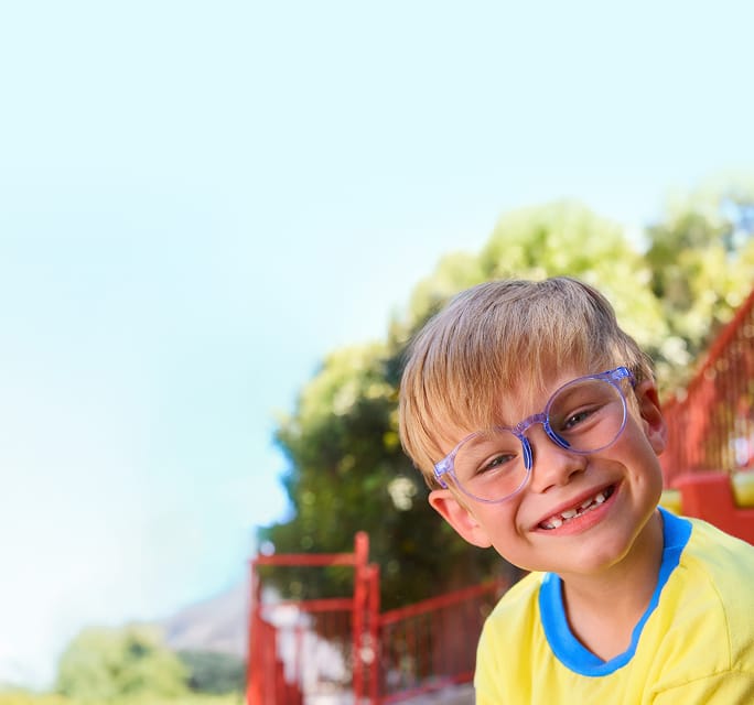 Young child with blonde hair and purple glasses smiling outdoors. He is dressed in a bright yellow shirt with a blue collar. The background is outdoors with the blurred outline of trees and a red stair railing, creating a colorful and cheerful setting.