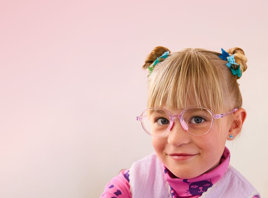 Child with pink translucent glasses and hair in buns, wearing a pink outfit with cat pattern, against a pink gradient background.
