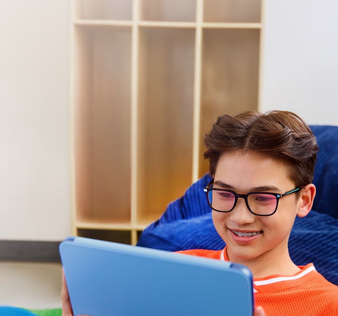 The image shows a young person with short brown hair, black rectangular glasses, and braces, smiling while looking at a blue tablet. They wear an orange shirt and sit on a blue cushion, with a blurred wall and bookshelf in the background.