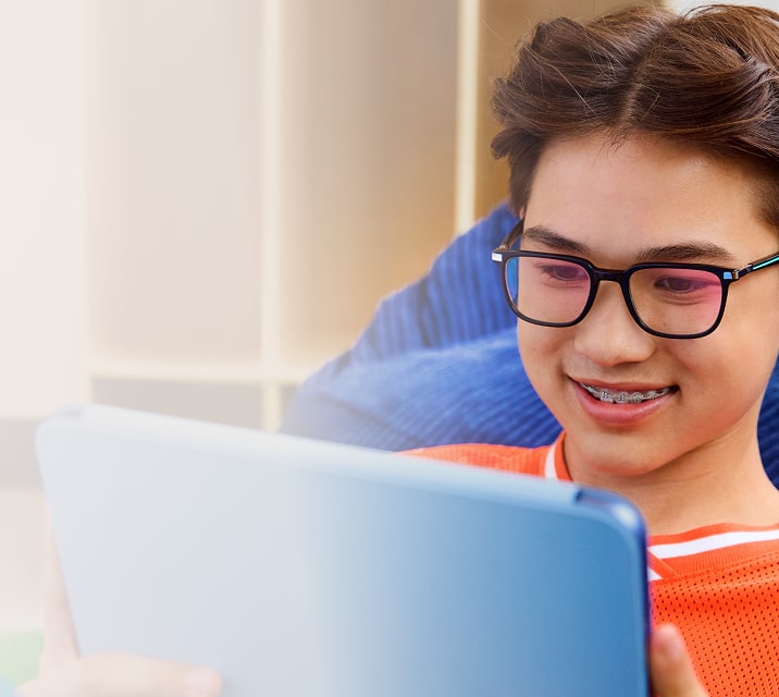 The image shows a young person with short brown hair, black rectangular glasses, and braces, smiling while looking at a blue tablet. They wear an orange shirt and sit on a blue cushion, with a blurred wall and bookshelf in the background.