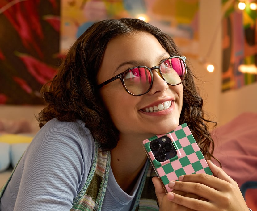 Teen girl holding a phone with checkered case, smiling while wearing glasses with EyeQLenz with Zenni ID Guard pink-tinted lenses indoors.
