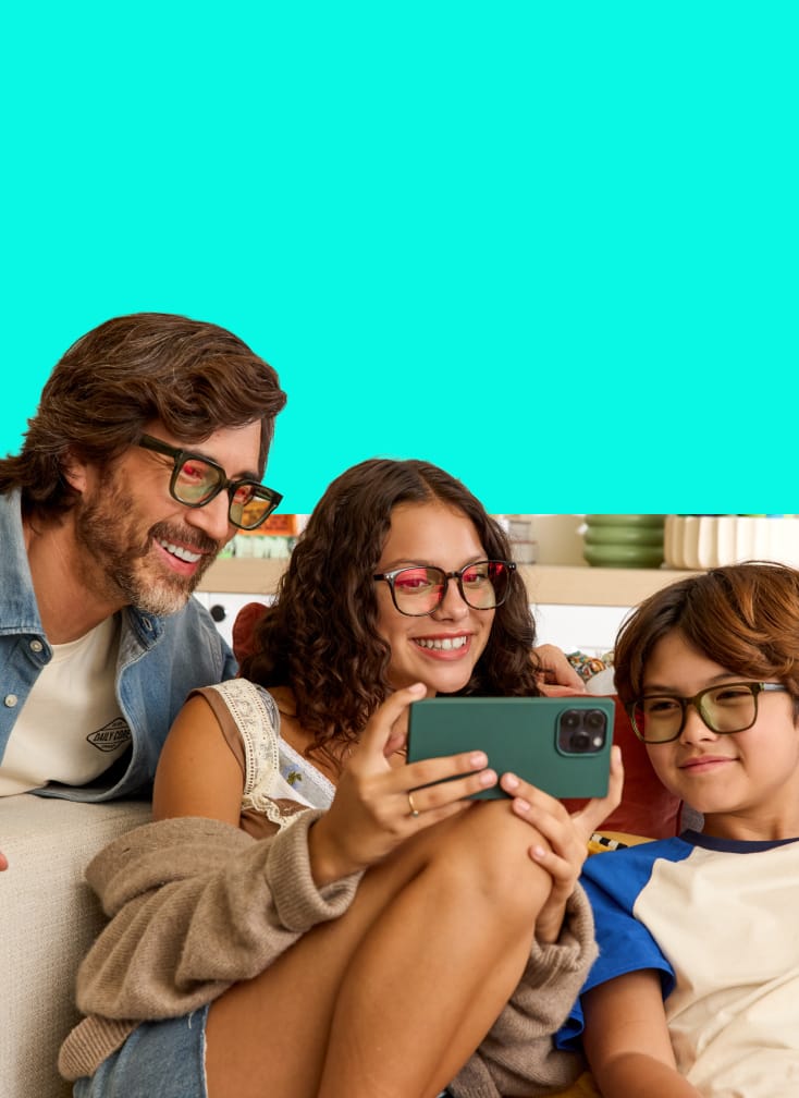 Man, teen girl, and child wearing EyeQLenz with Zenni ID Guard glasses and smiling while looking at a phone together on the couch.