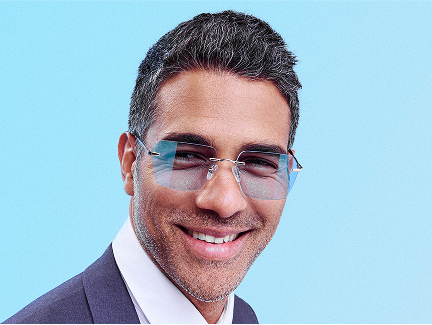 A man with short curly hair and a beard wearing golden-brown eyeglasses. He is smiling brightly and dressed in a burgundy and white patterned collared shirt, on a light blue background with a foliage motif.