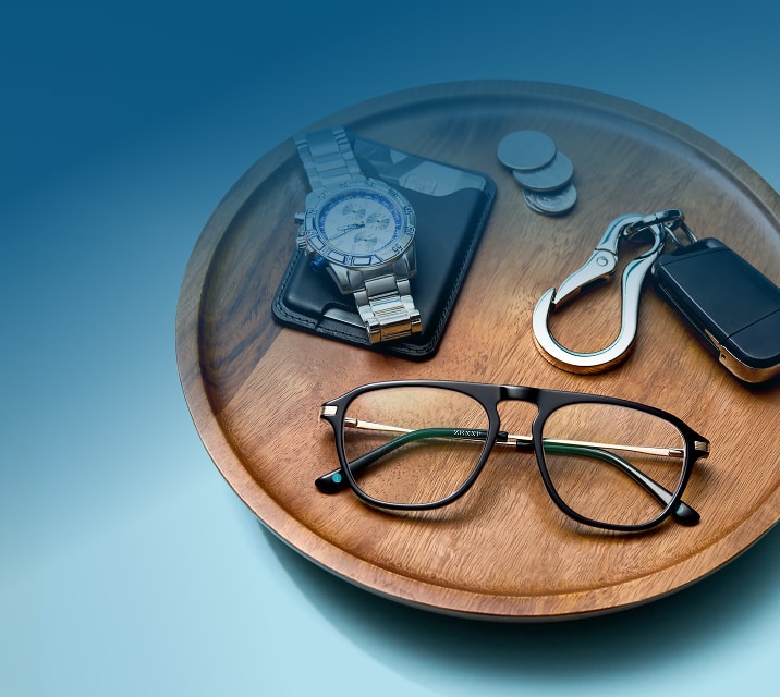 Black aviator-style eyeglasses with gold metal temples resting on a round wooden tray alongside a silver watch, leather wallet, coins, and a keychain with a carabiner.