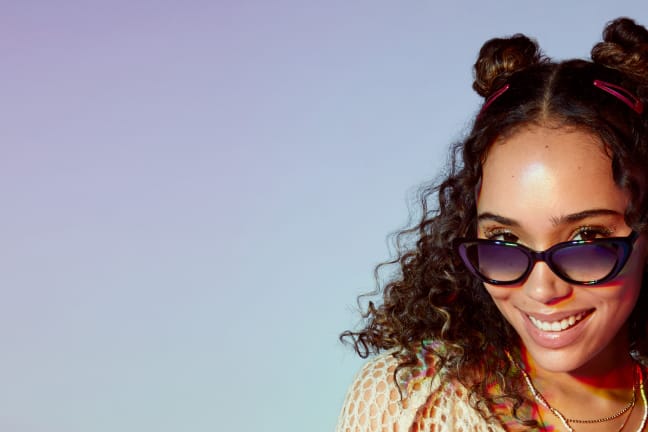 A cheerful woman with curly hair and rimless Zenni glasses poses against a pastel background, embodying the festive spirit.