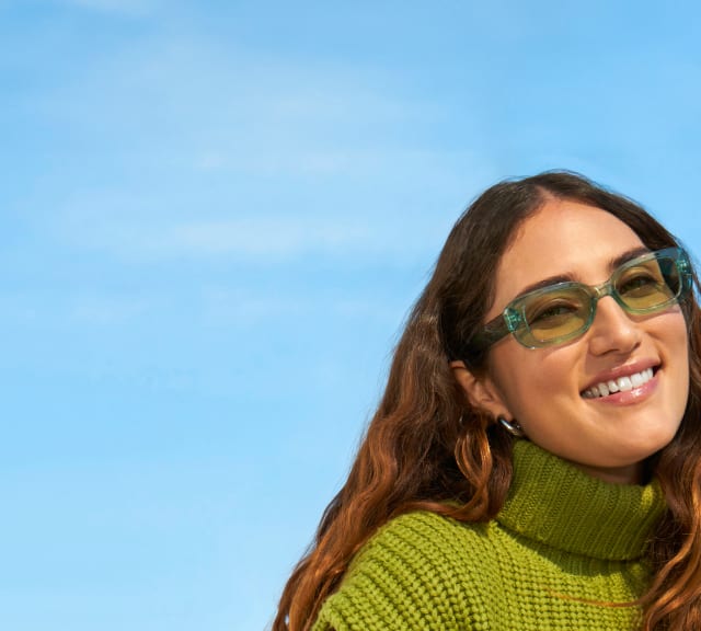  A woman with long wavy brown hair smiles while wearing green-tinted sunglasses. She is dressed in a textured green turtleneck sweater, with a blue sky as the backdrop.