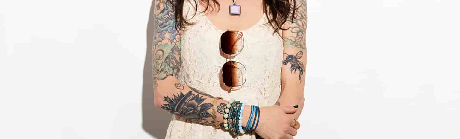 Woman with face cropped out shows off her arm tattoos in a  white lace sleeveless dress with oversized glam sunglasses #1120814 hanging on the neckline.