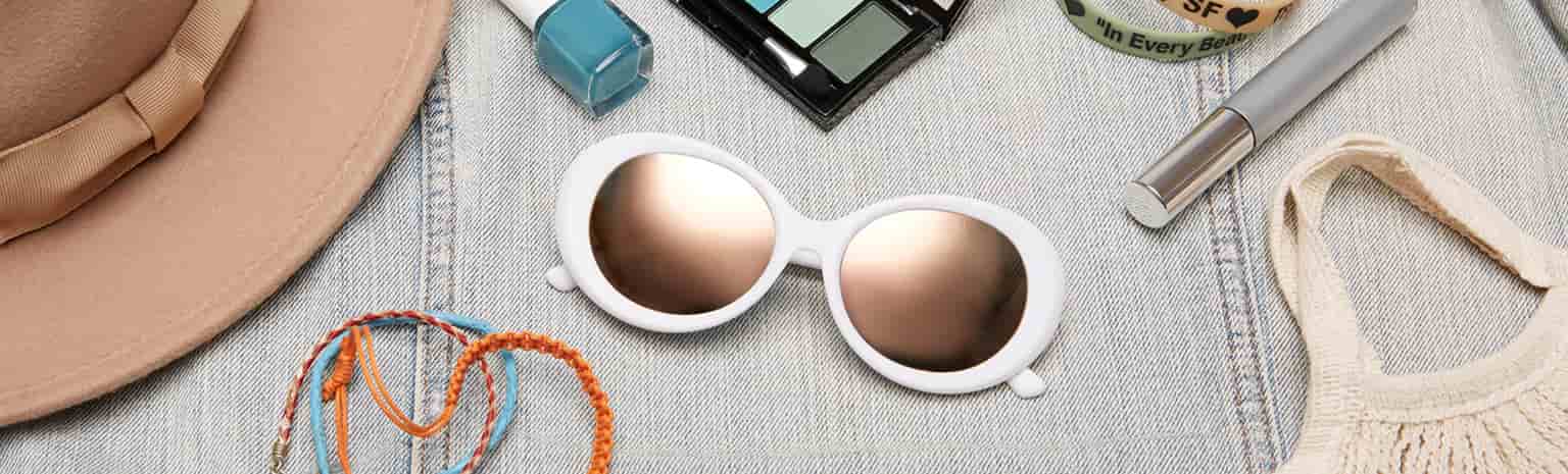White oval sunglasses #2024730 surrounded by beige fedora, blue nail polish, blue and green eyeshadow palette, string bracelets, plastic wristbands, and boho macramé bag.