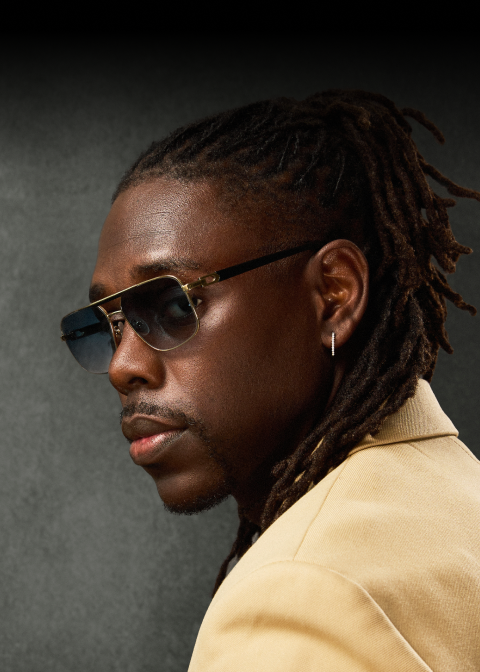 Boston Celtics point guard Jrue Holiday wears “TripleDouble” aviator sunglasses from his collection with Zenni while looking over his shoulder at the camera wearing a beige jacket accessorized with silver earrings in front of a dark backdrop.