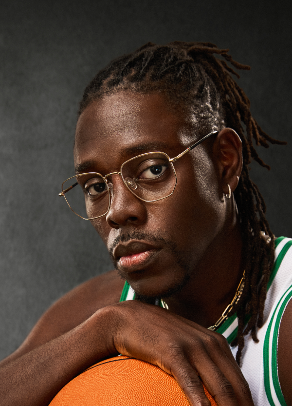 Boston Celtics point guard Jrue Holiday is pictured wearing gold “Hendrix” optical glasses from his collection with Zenni, staring directly into the camera, holding a basketball, dressed in his NBA jersey against a dark backdrop.