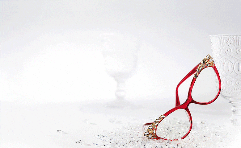 Zenni Red-framed cat-eye glasses with decorative crystals resting on a frosty white surface beside a crystal glass. Zenni sport sunglasses with mirrored lenses on top of snow mound next to a pine cone and evergreen sprig. Zenni sunglasses propped up in beach sand beside a small sandcastle and a blue sky in the background.