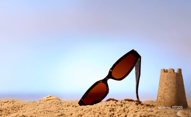 Zenni sunglasses propped up in beach sand beside a small sandcastle and a blue sky in the background.