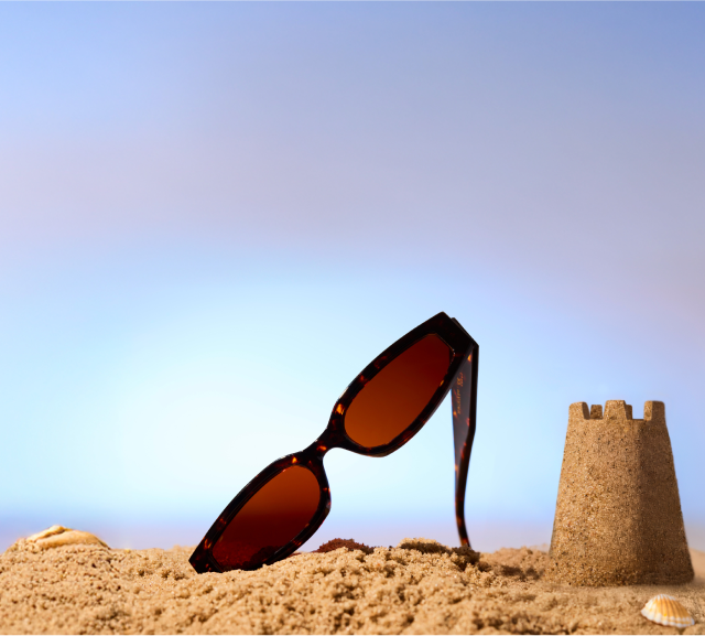 Zenni sunglasses propped up in beach sand beside a small sandcastle and a blue sky in the background.