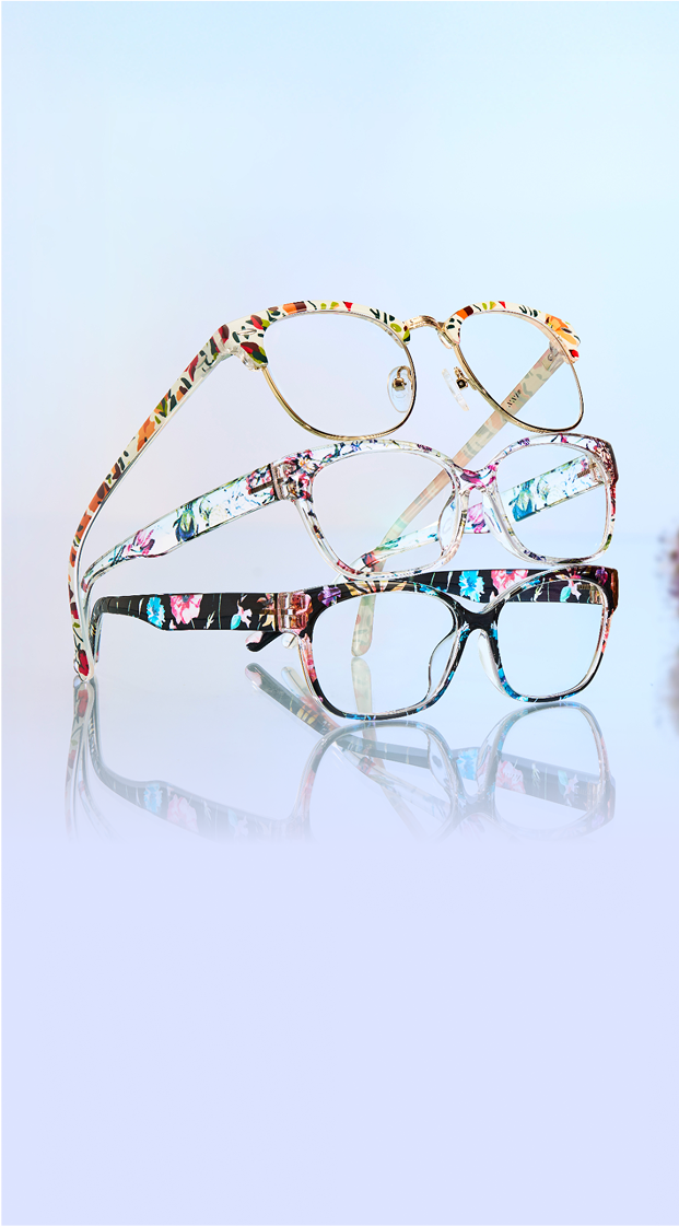 A stack of three floral-patterned eyeglasses is displayed against a soft blue background, with their reflections on a glossy surface and blurred flowers in the background.