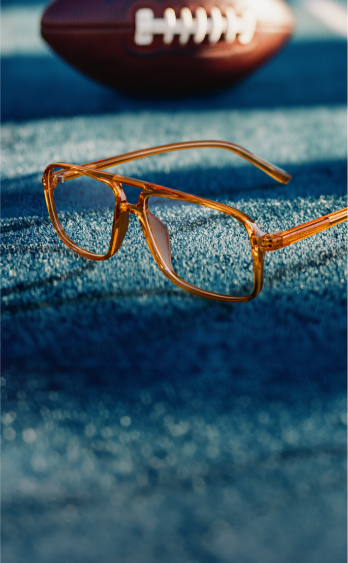 A pair of amber-colored eyeglasses rests on a blue surface, with a blurred American football in the background.