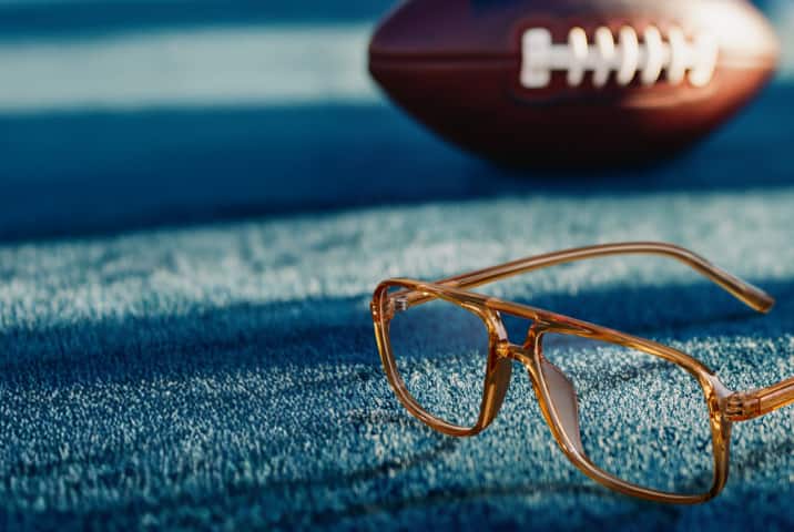 Amber-colored eyeglasses resting on a blue textured surface resembling a football field, with a blurred football in the background.