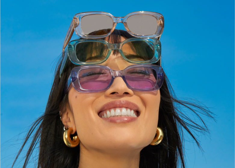 Smiling woman wearing three pairs of sunglasses stacked on her head, against a clear blue sky.