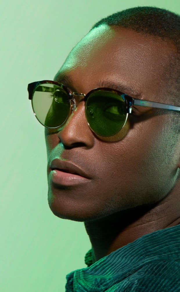A man with short buzzed hair wearing browline glasses with green lenses.