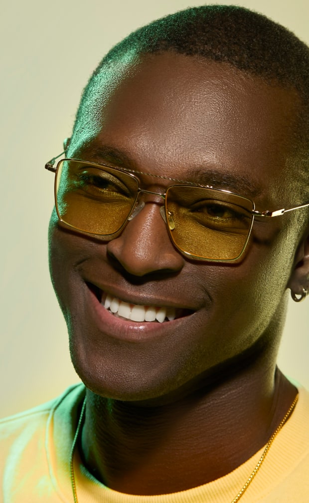 A smiling man with short buzzed hair wearing gold metal aviator glasses with yellow lenses.