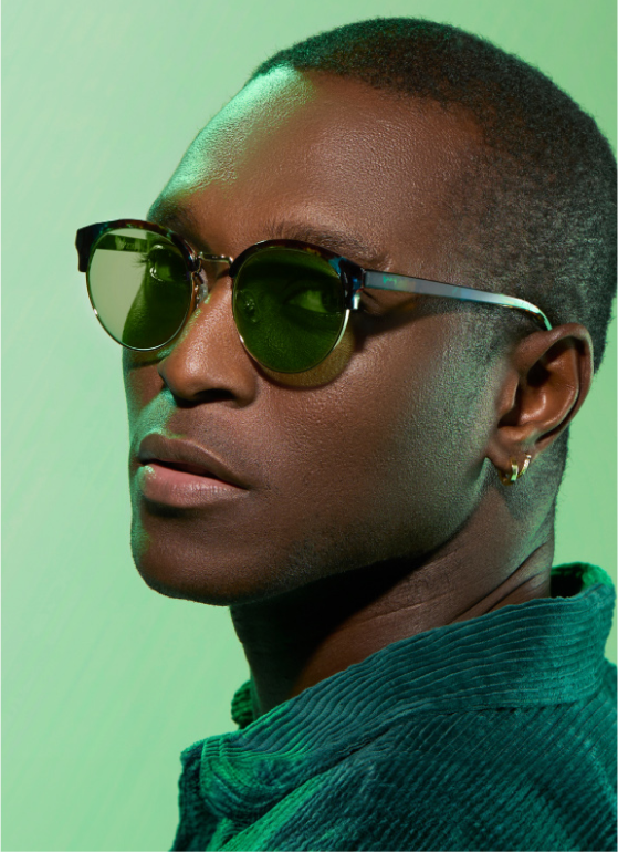 A man with short buzzed hair wearing browline glasses with green lenses.