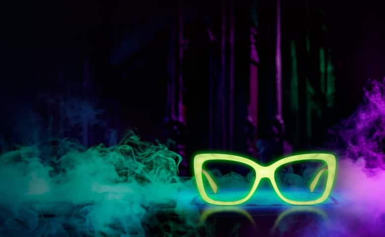 The image features a pair of neon green, cat-eye style glasses glowing against a dark background. Surrounding the glasses are swirling clouds of green and purple smoke, creating a mysterious and ethereal atmosphere.