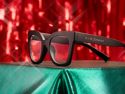 Black Zenni eyeglasses with ‘A & M Forever’ custom engraving on temple arm against festive red and green holiday lights background.